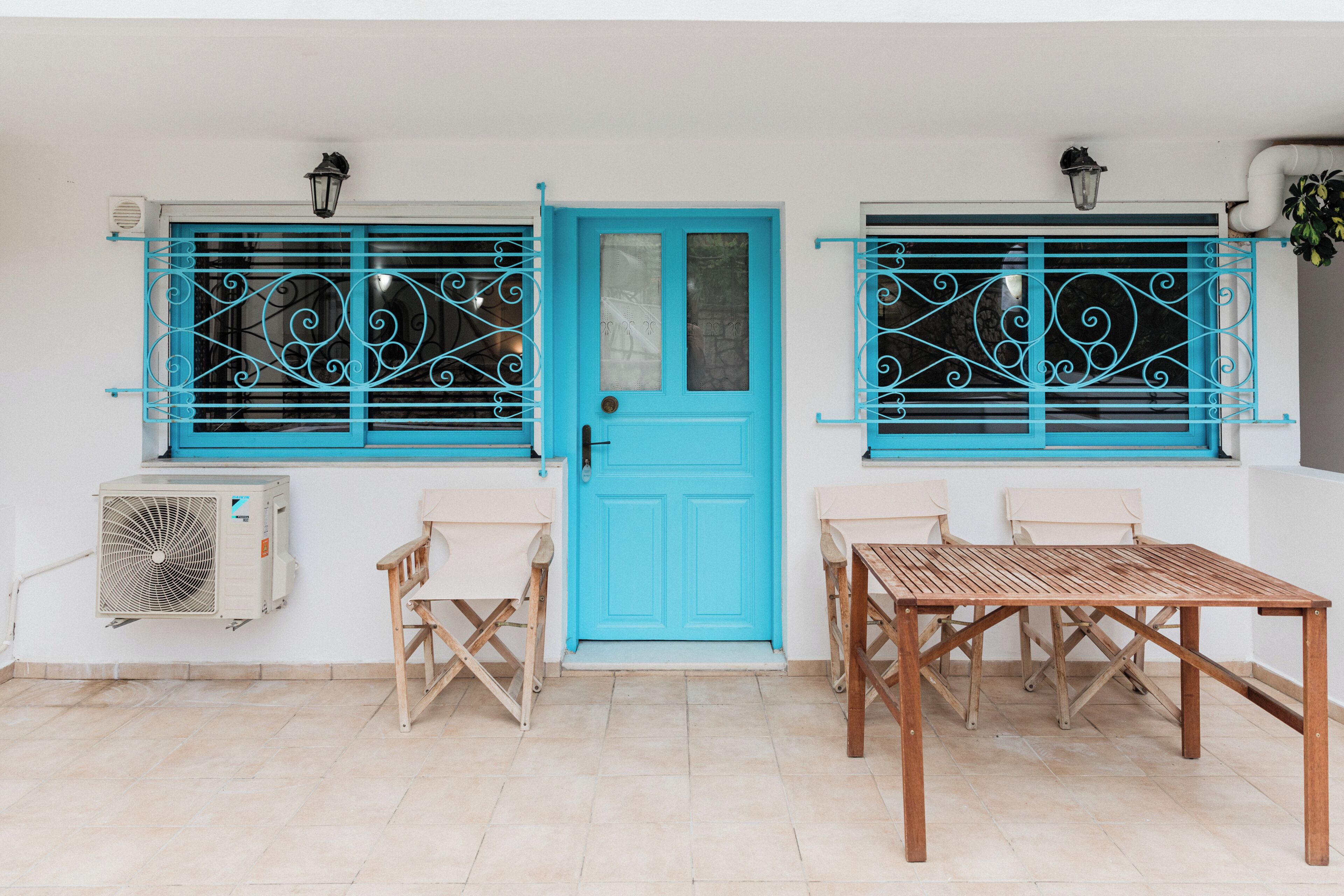 Una puerta azul, dos ventanas con celosías metálicas decorativas, una mesa de madera y dos sillas.