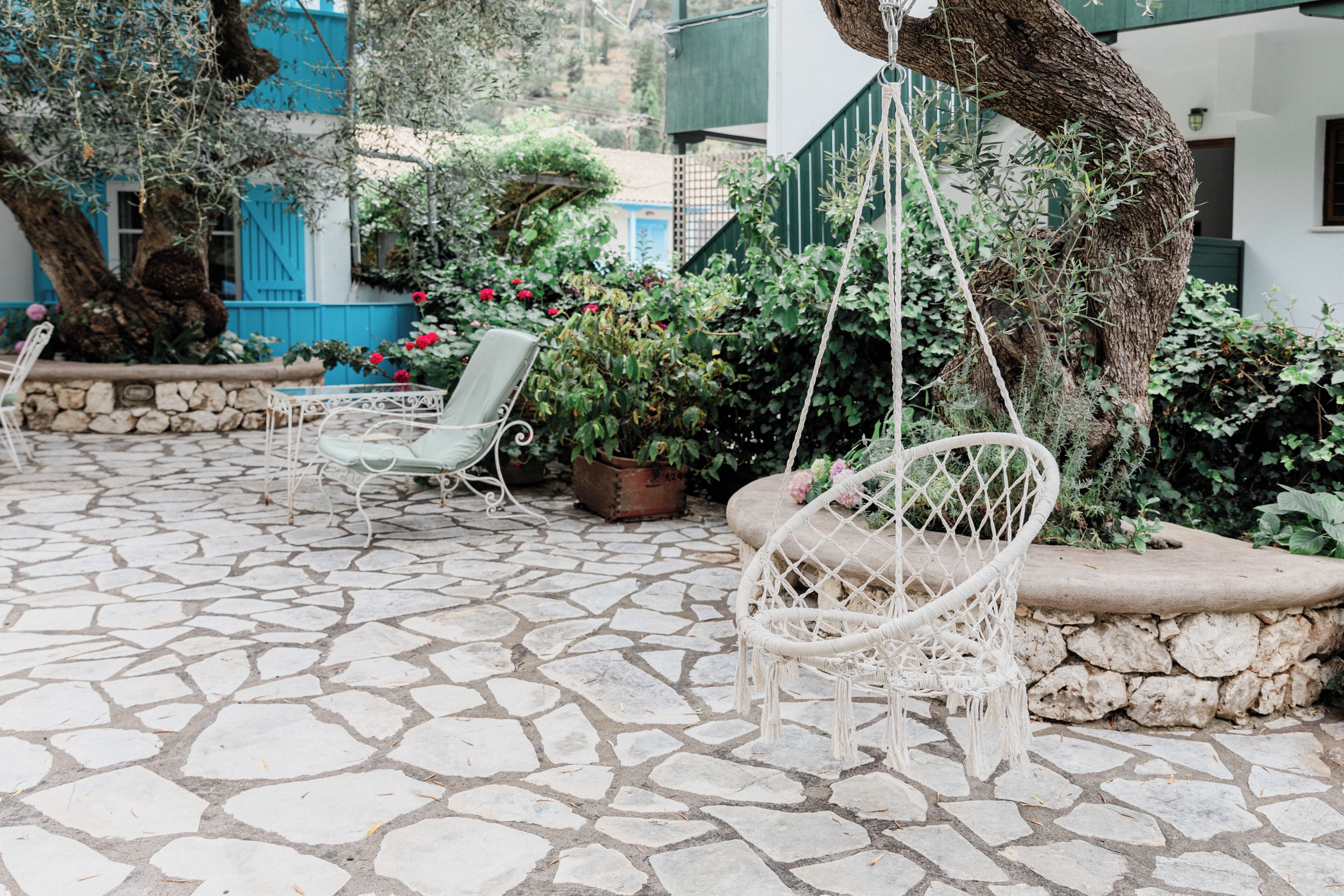 A stone-paved patio with two white wicker chairs, a hanging swing, and potted plants.