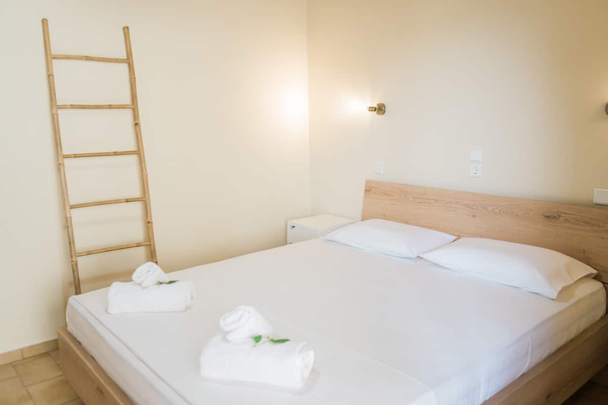 A neatly made bed with white linens and two rolled towels, a wooden headboard, a wall-mounted light, and a wooden ladder leaning against the wall.