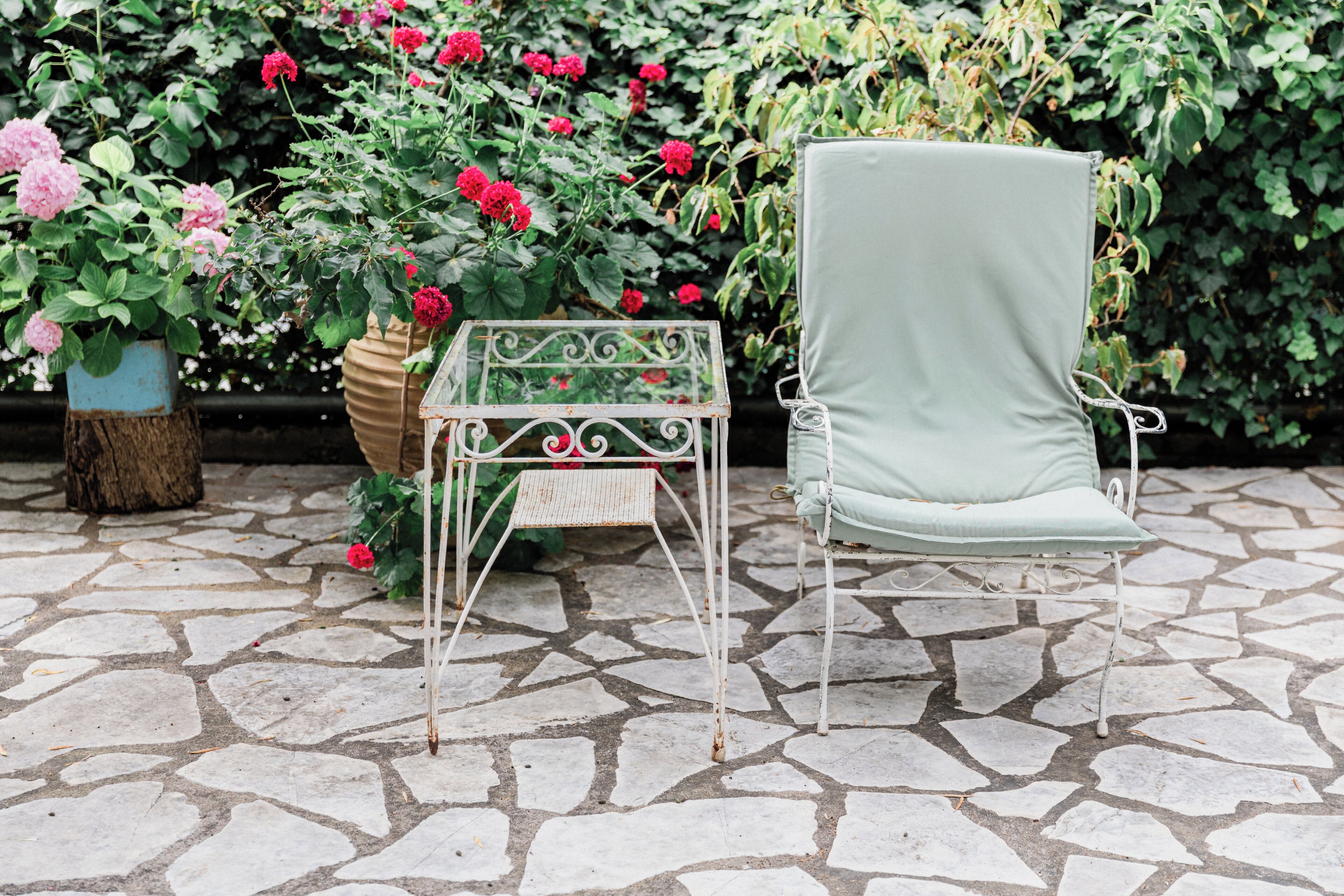 Un jardín con mesa y silla de metal, rodeado de plantas y flores en macetas.