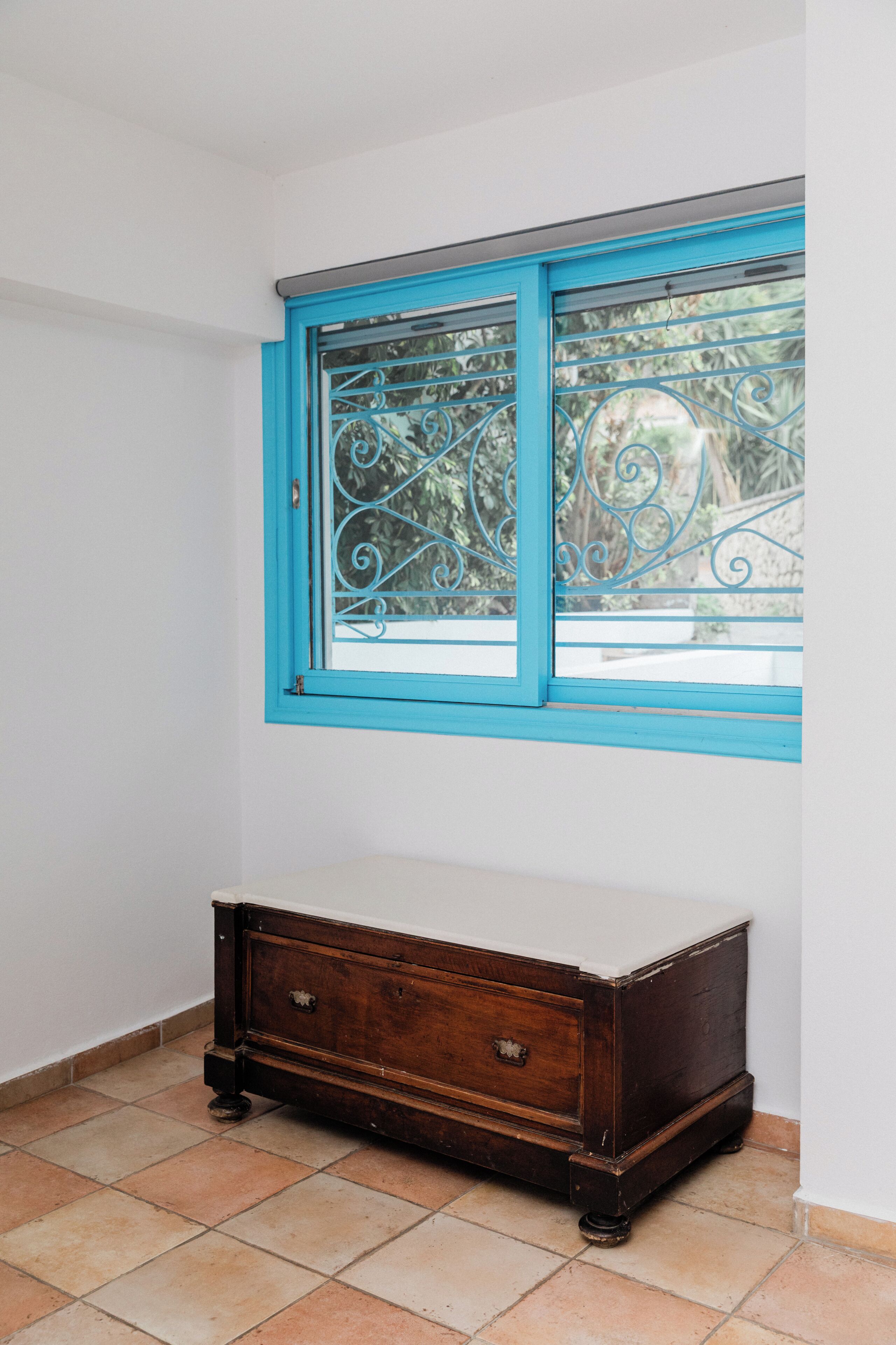 A room with a window with blue trim, a wooden dresser, and tiled flooring.