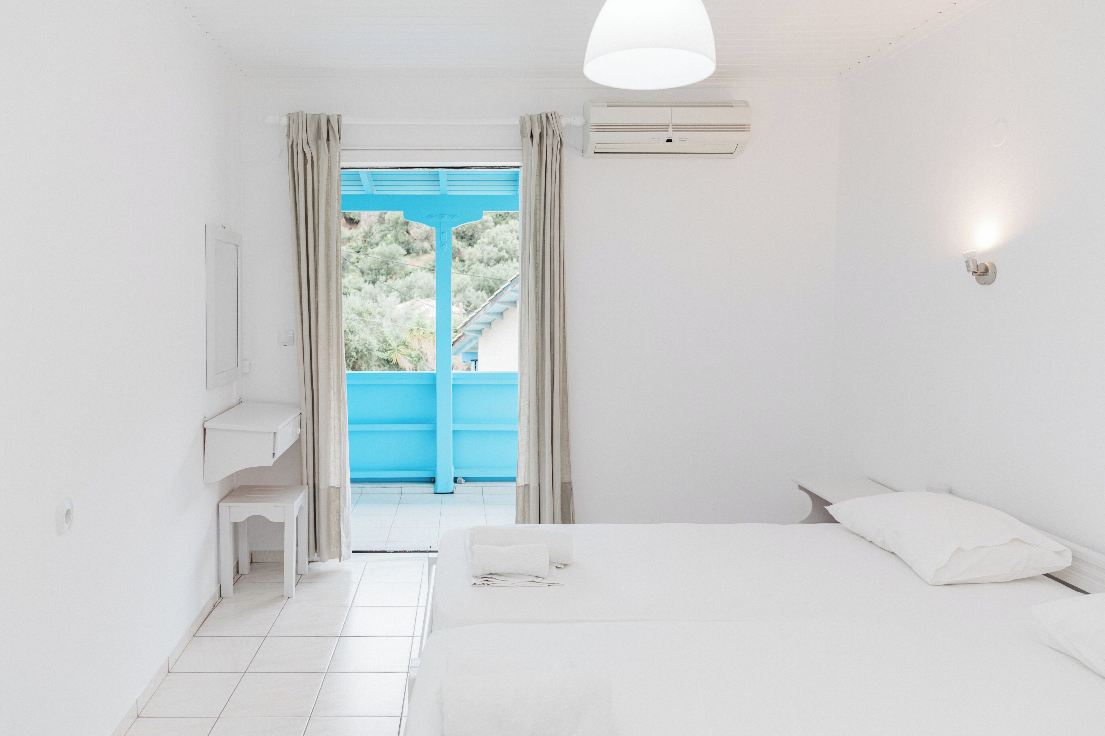 Ein helles Zimmer mit einem Bett, einem kleinen Tisch, einem Fenster mit Vorhängen und einer Klimaanlage.