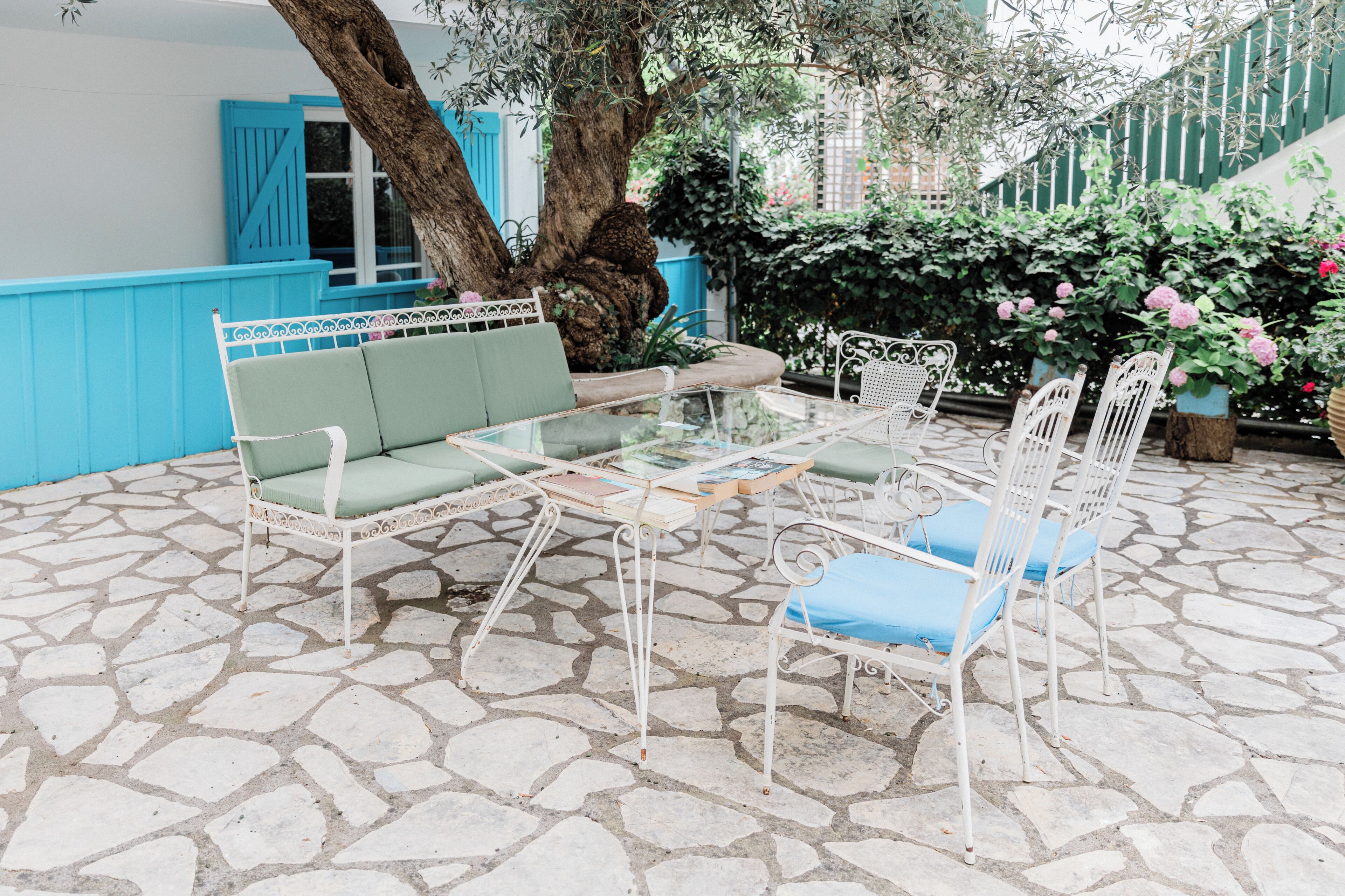 A stone-tiled patio with a glass-top table, white metal chairs, and a green cushioned bench.