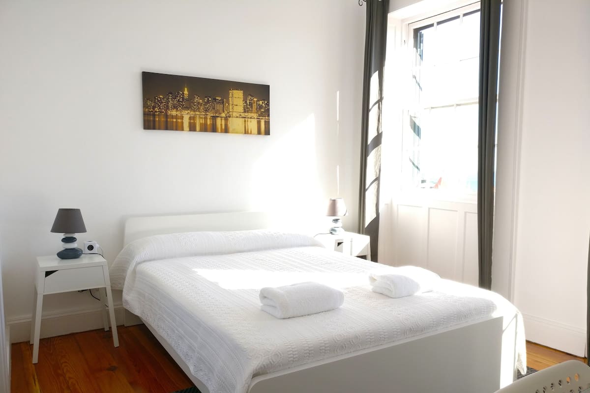 Un lit bien fait, avec du linge de lit blanc et deux serviettes, une petite table de chevet avec une lampe, un tableau représentant un paysage urbain encadré au mur, et une fenêtre avec des rideaux.