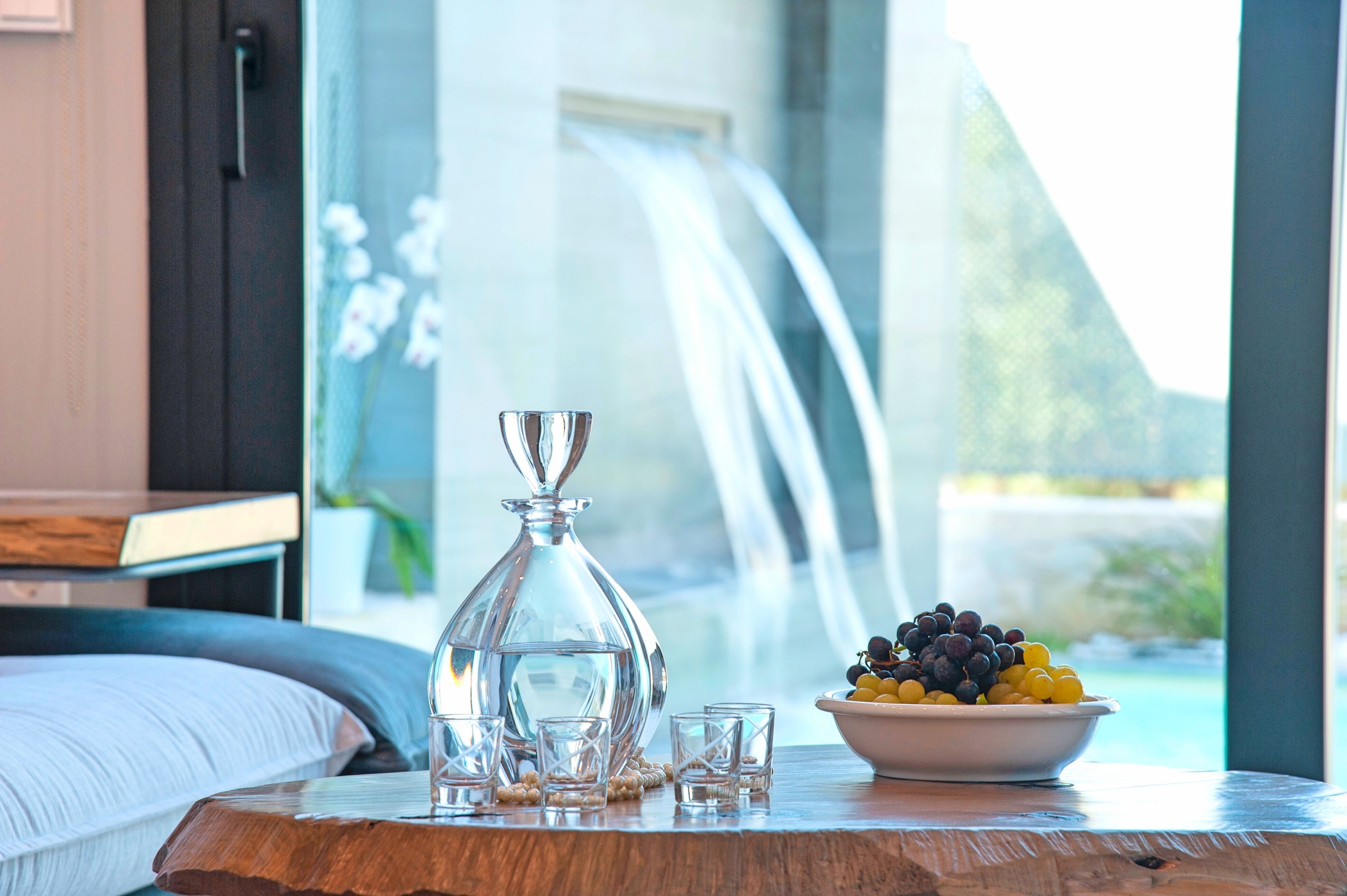 A bedroom with a bed, a wooden table with a glass decanter, glasses, and a bowl of fruit. A waterfall is visible through the window.