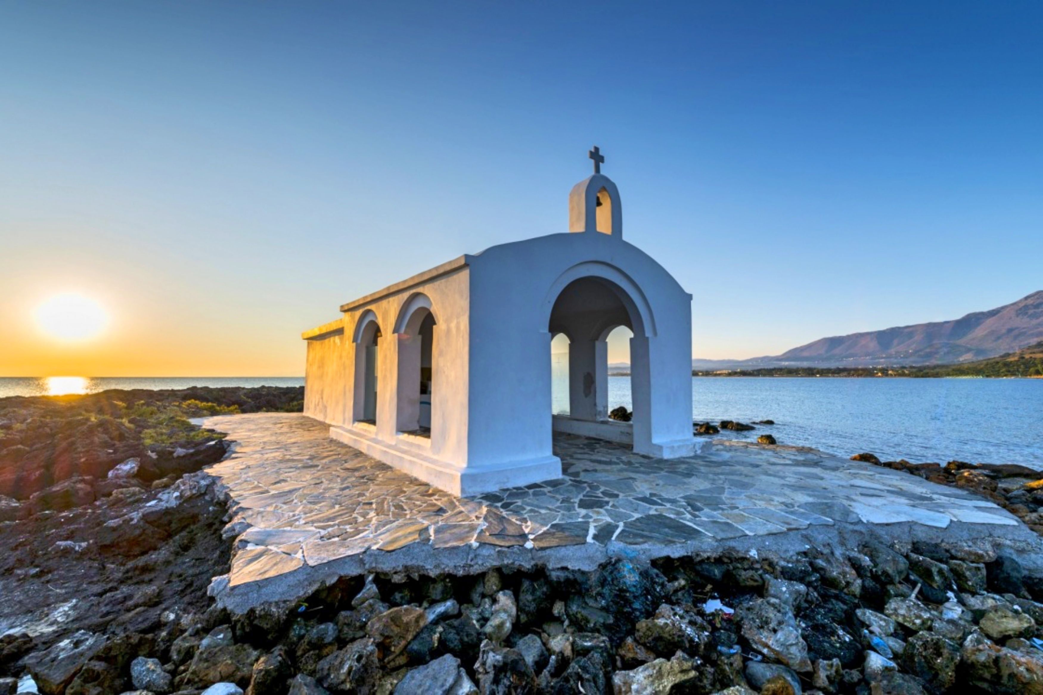 A white church with a cross on top, situated on a rocky shore with a view of the sea and mountains in the background.