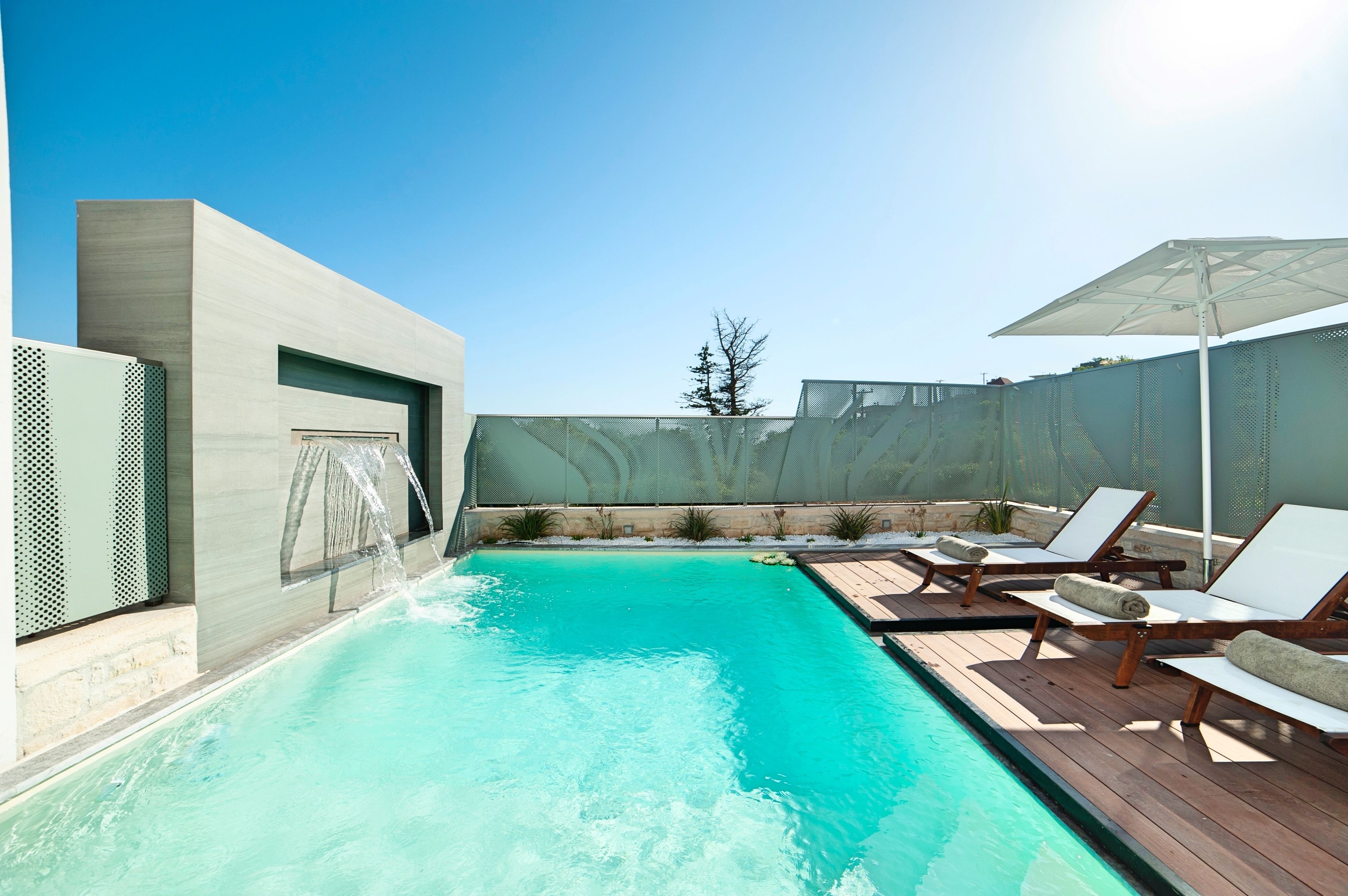Piscina com espreguiçadeiras e um guarda-sol, cercada por um edifício moderno com grade de metal.