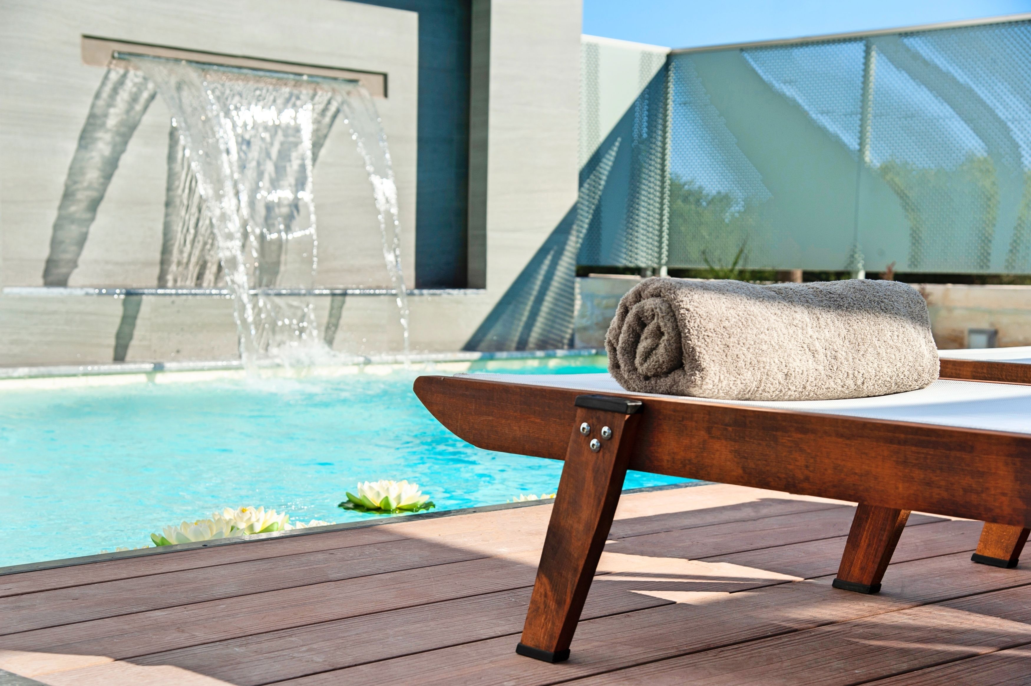 A wooden lounge chair with a towel, a pool with a waterfall feature, and a modern building with a mesh screen in the background.