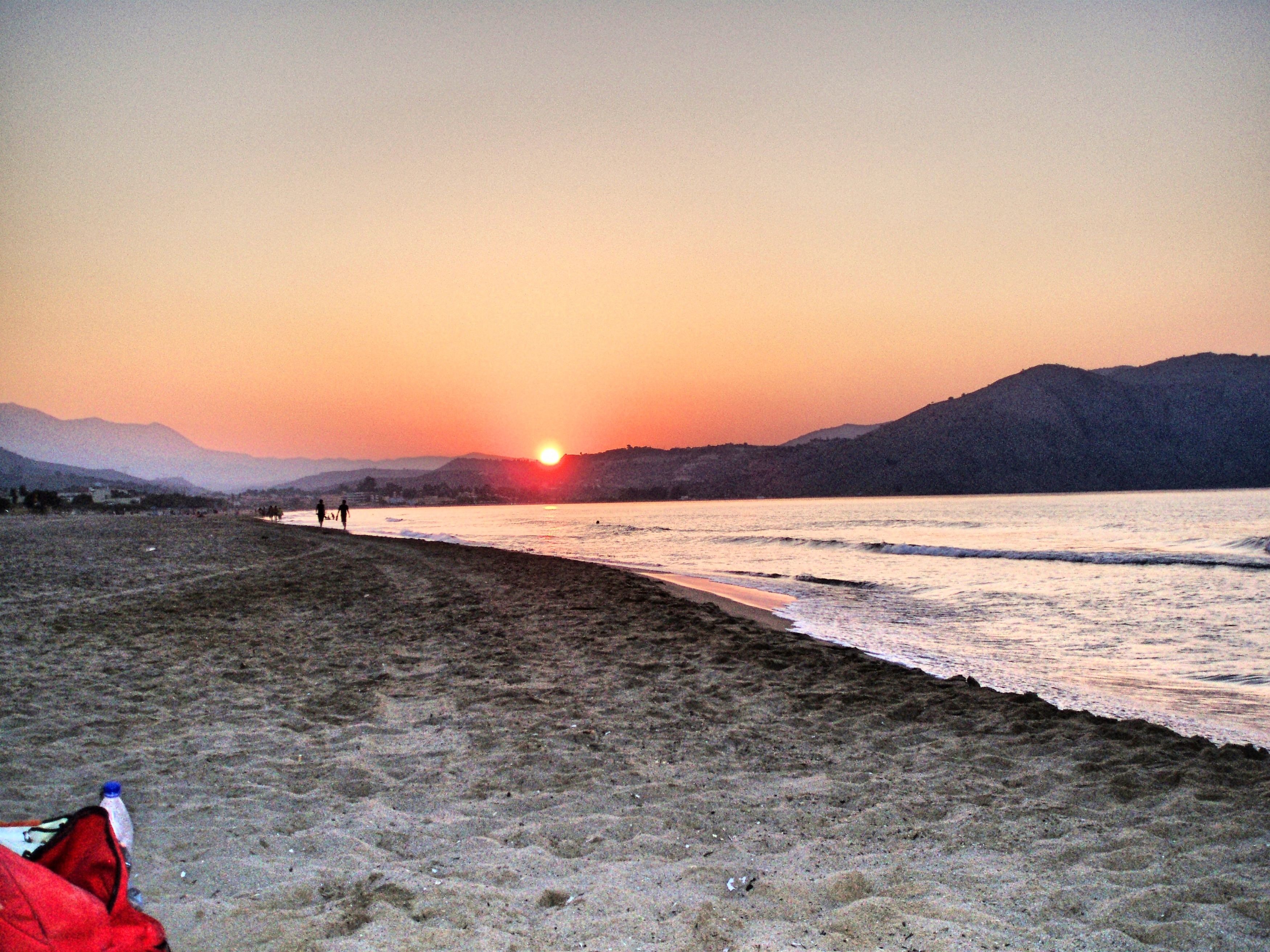 A beach at sunset with a clear sky, calm water, and distant mountains.