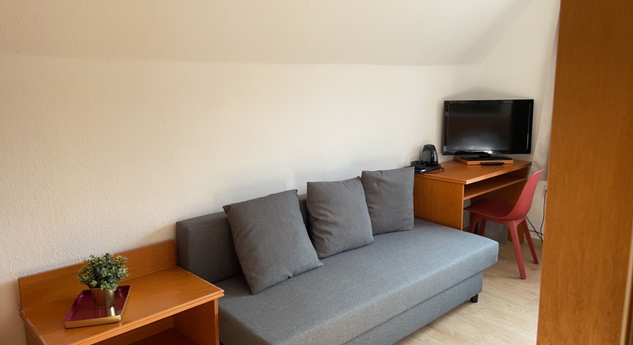 Una habitación pequeña y moderna con un sofá gris, un escritorio de madera, un televisor y una silla roja.
