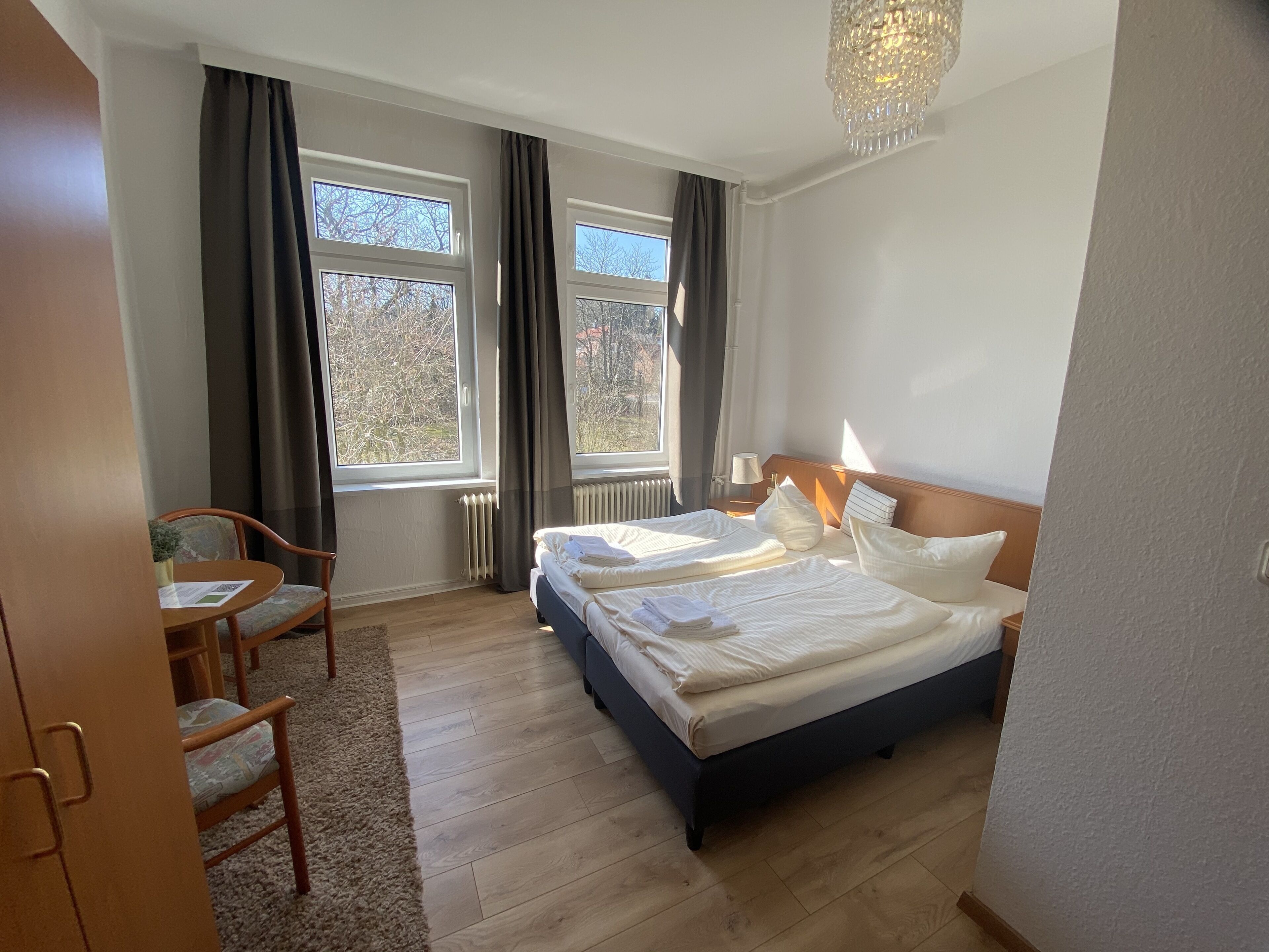 A hotel room with a bed, two windows, a chair, a table, and a chandelier.
