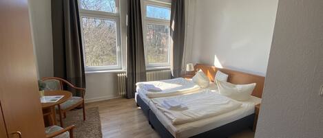 A hotel room with a bed, two windows, a chair, a table, and a chandelier.
