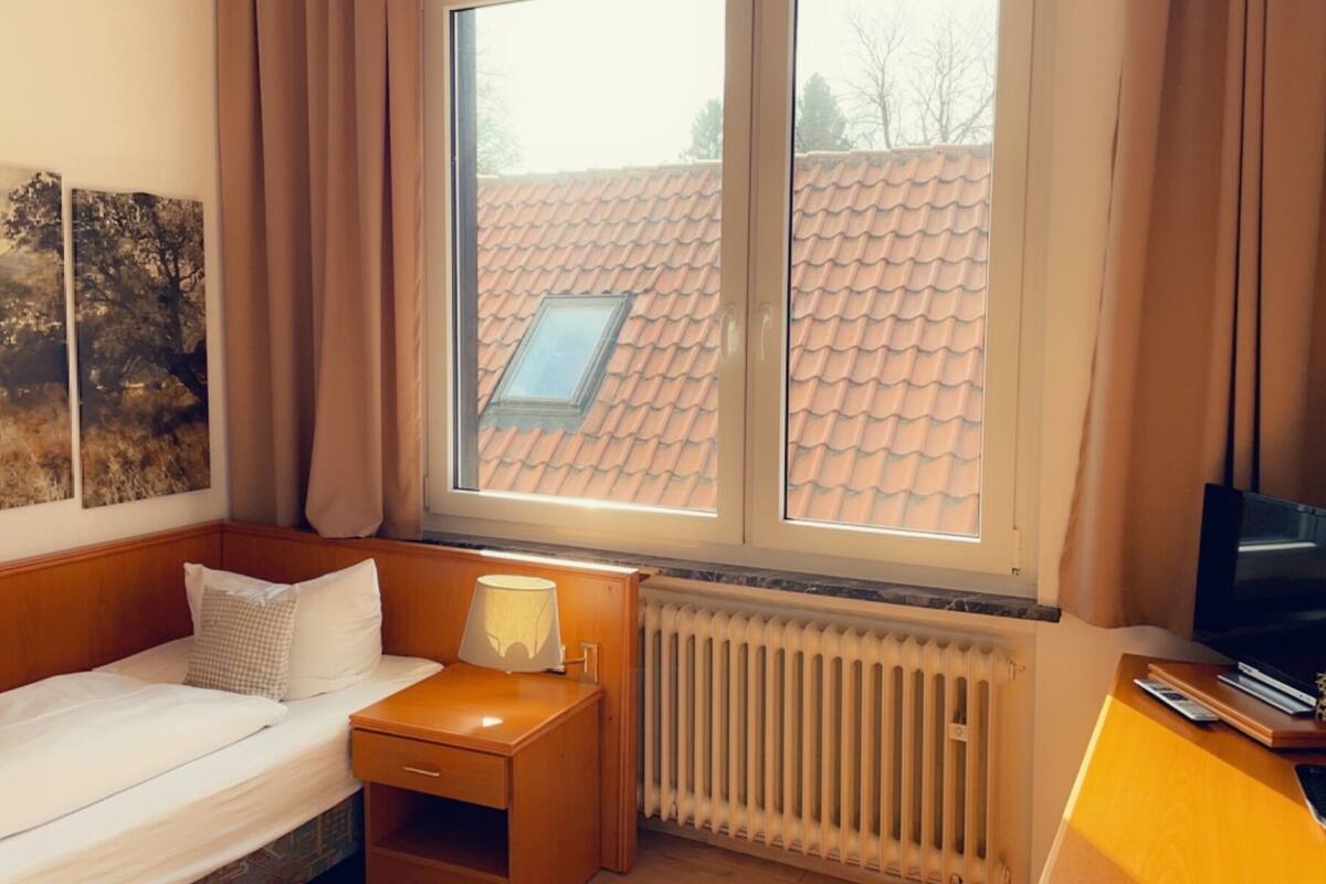 A hotel room with a bed, a desk, a radiator, and a window with a view of a tiled roof.