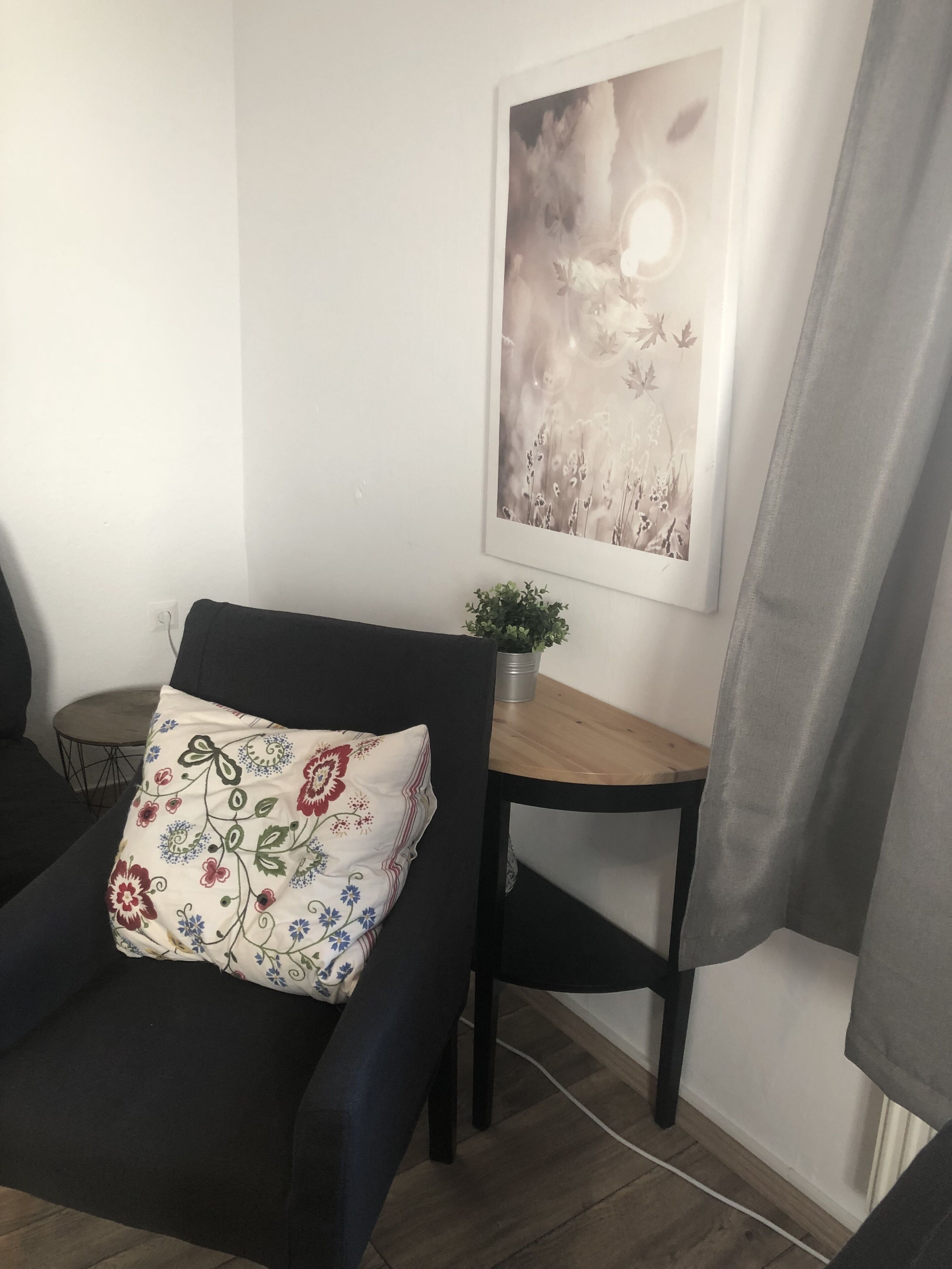 Uma sala de estar moderna com uma poltrona preta, uma pequena mesa de madeira, um vaso com planta e uma obra de arte floral emoldurada na parede.