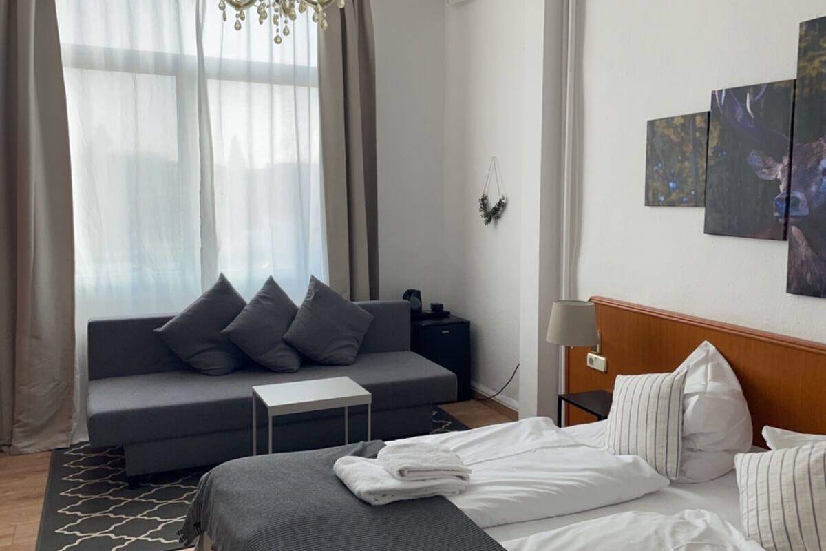 A hotel room with a bed, a grey sofa, a small table, a nightstand, and a wall with three framed pictures.