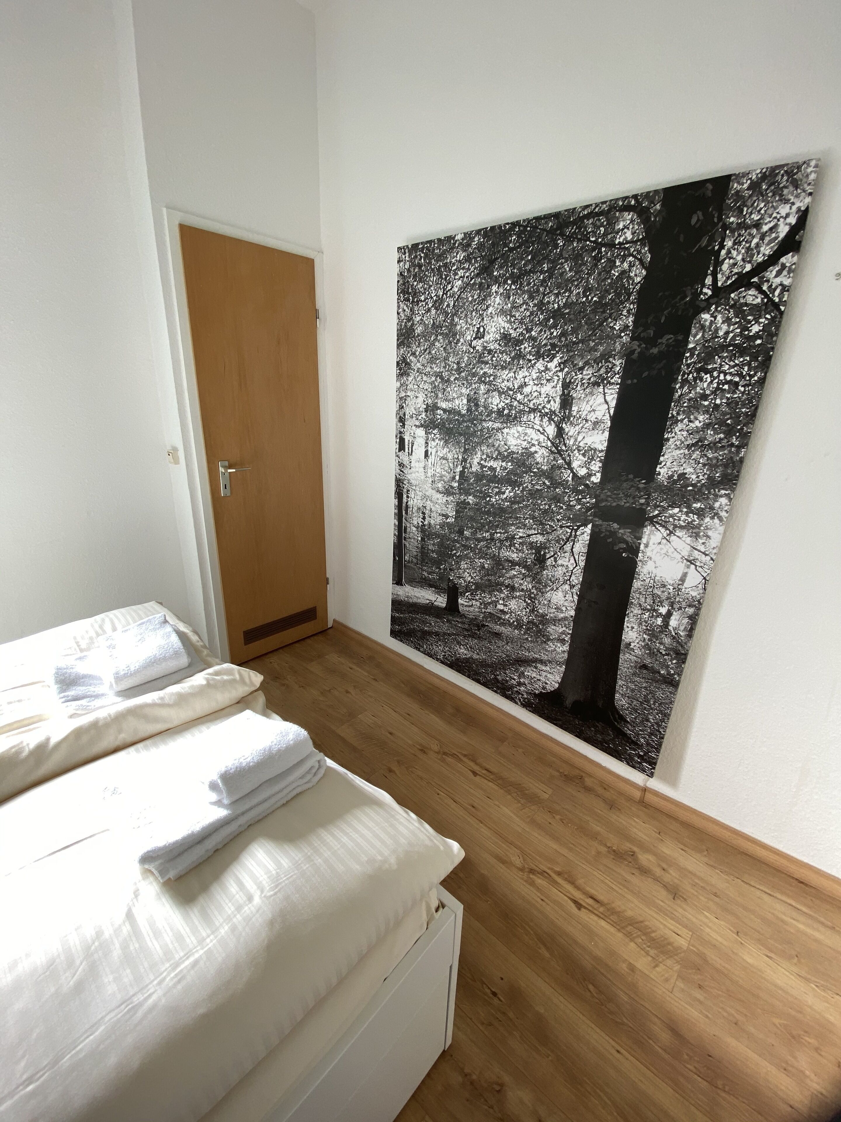 A bedroom with a wooden floor, a bed with white linens, and a large black and white forest photograph on the wall.