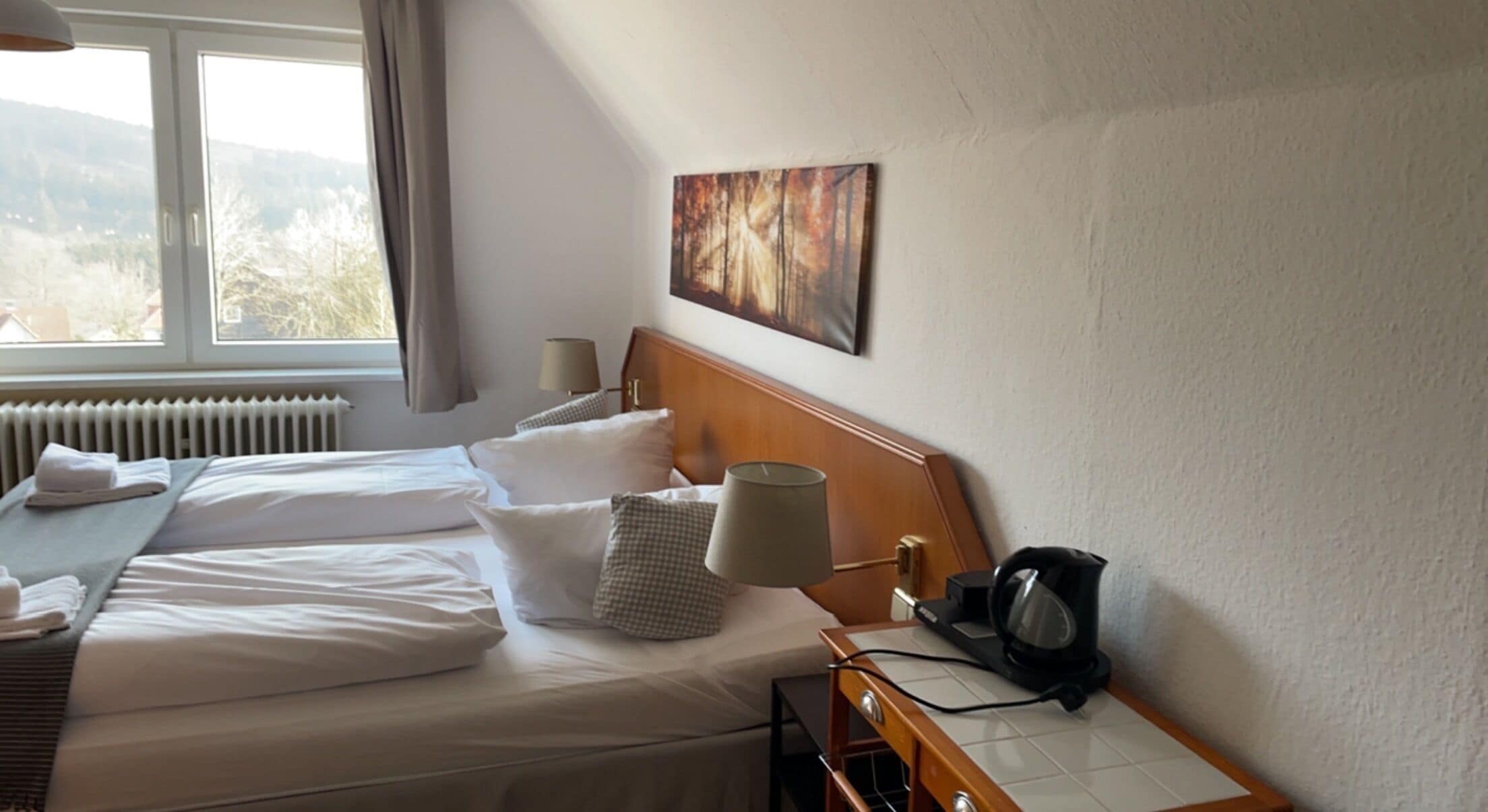 A hotel room with two beds, a radiator, a painting, a lamp, and a kettle on a table.