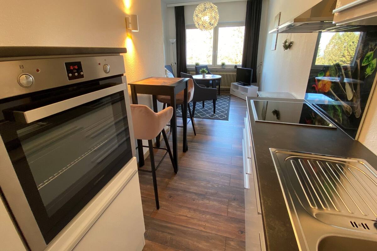 A compact kitchen with a built-in oven, a stainless steel range hood, and a dining area with a table and chairs.