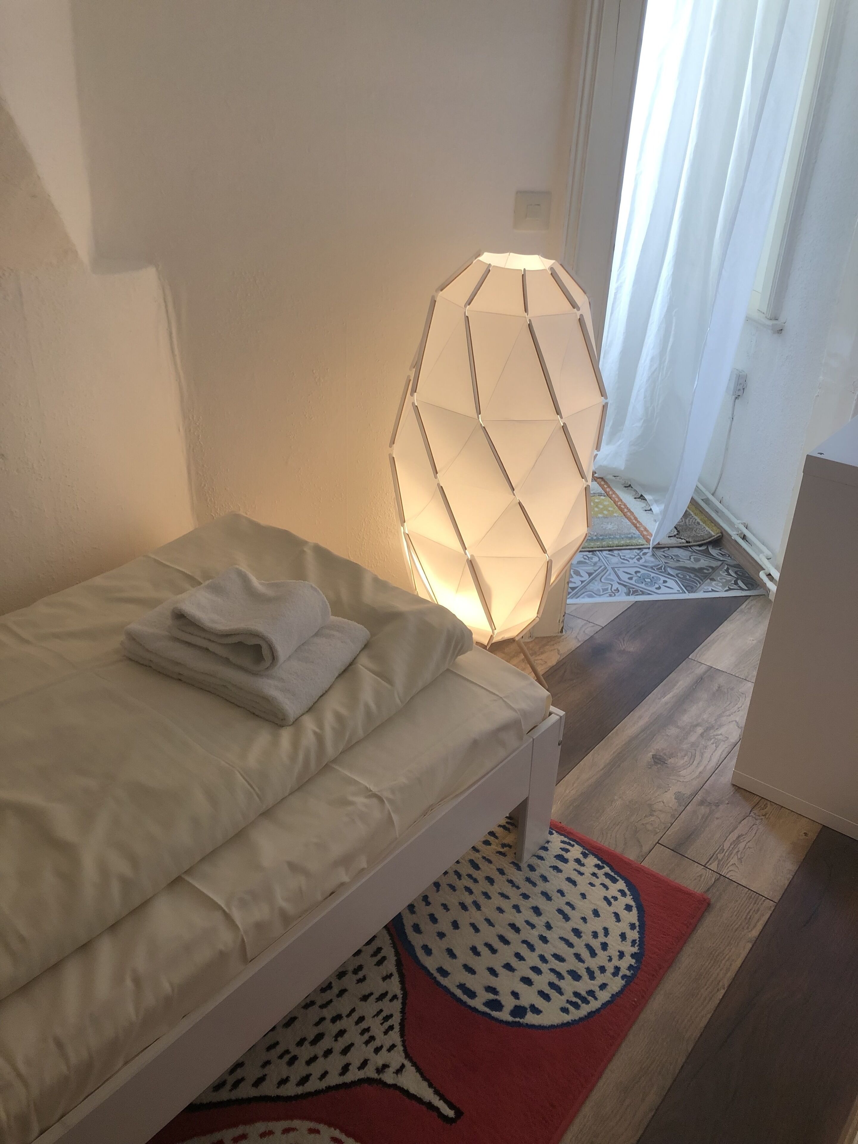 A neatly made bed with a folded towel, a geometric-patterned lamp, and a patterned rug on a wooden floor.