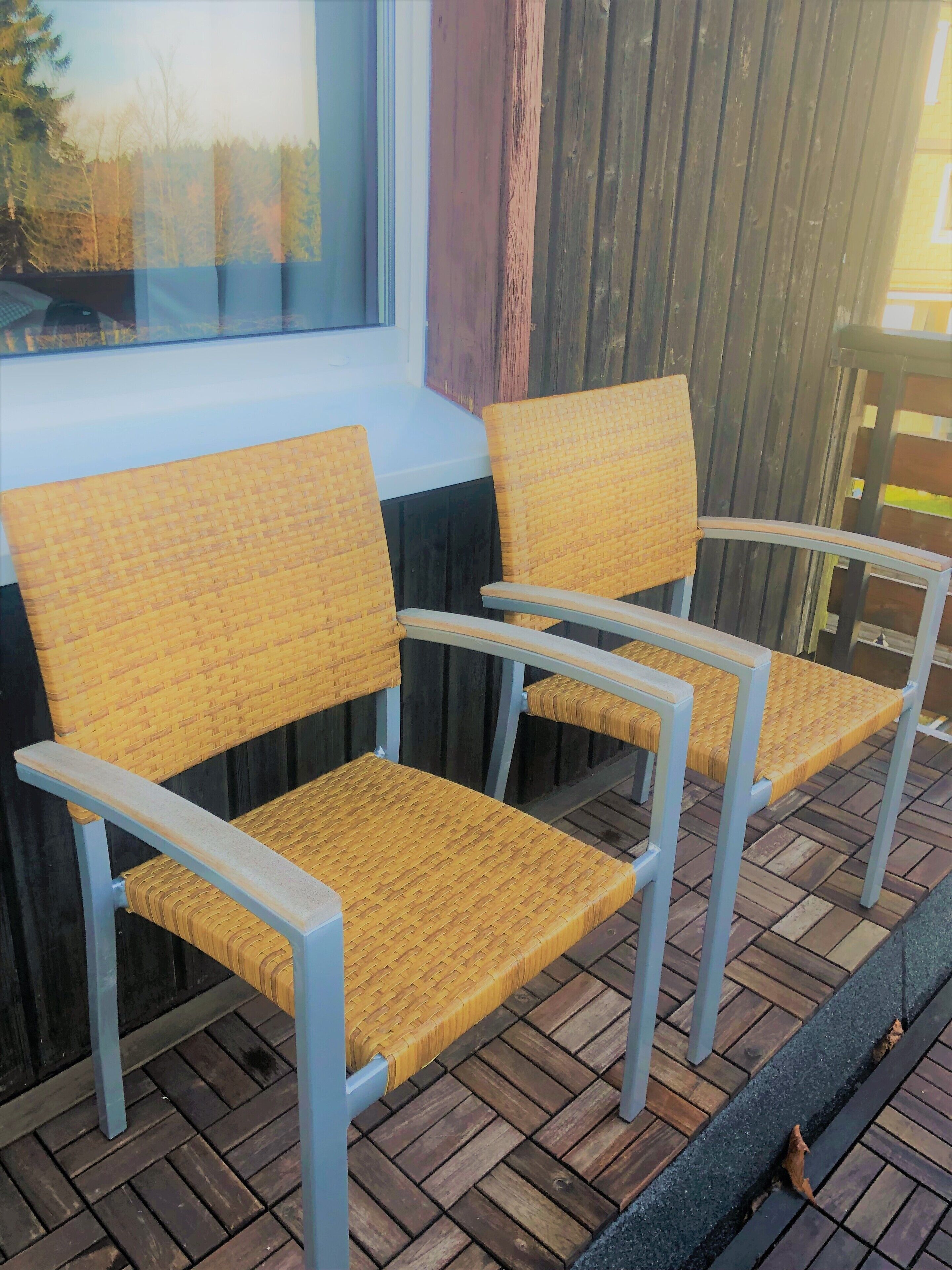 A set of four wicker chairs with metal frames, arranged on a wooden deck with a window in the background.