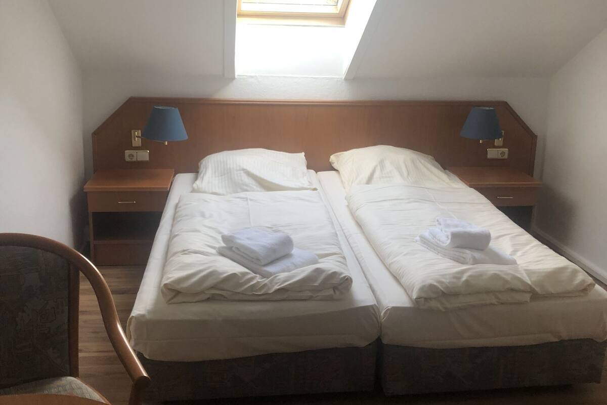 A room with two beds, a skylight, a wooden nightstand with a lamp, and a chair.