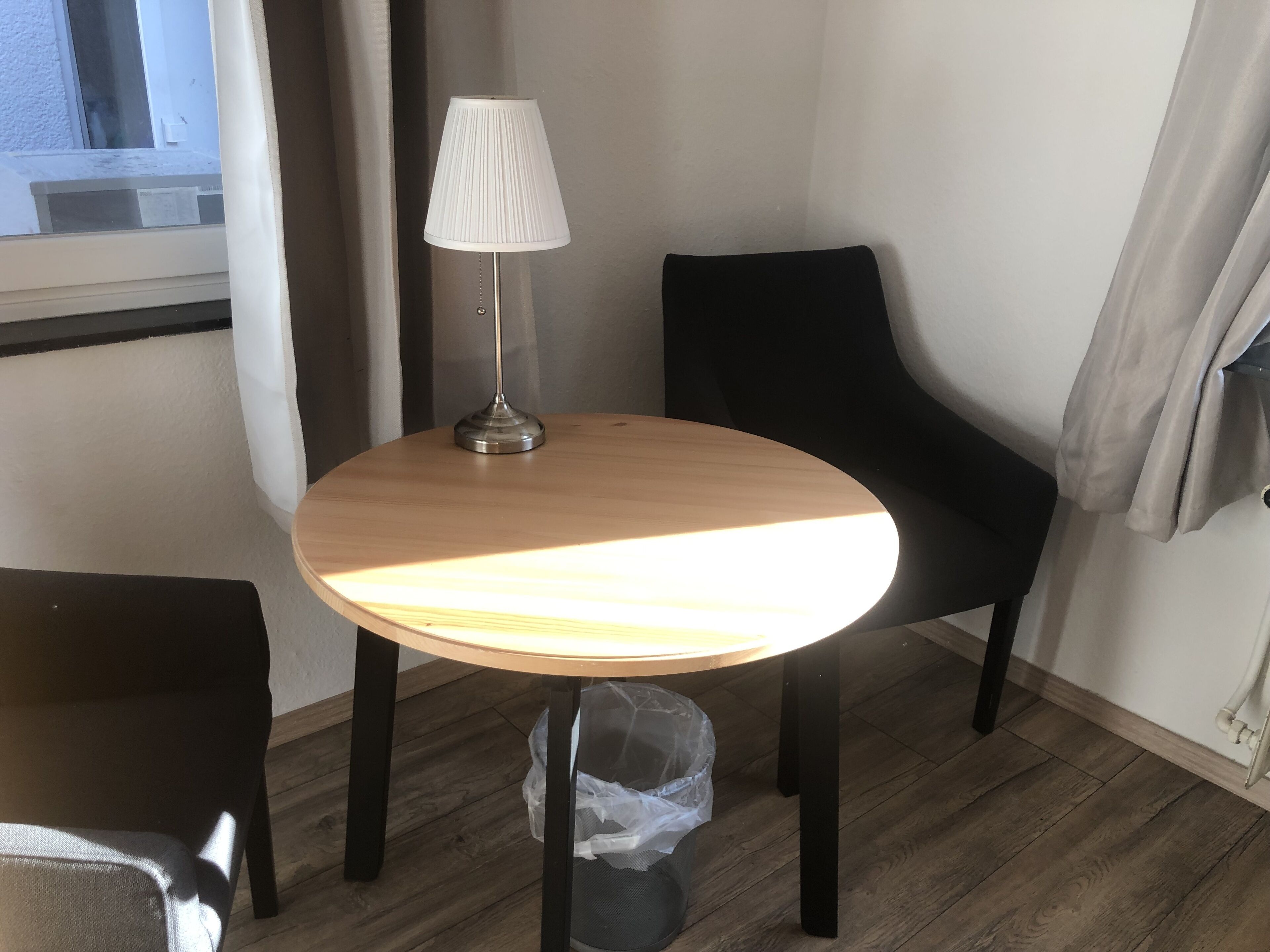 Una mesa redonda de madera con una pantalla blanca para lámpara, una silla negra y un cubo de basura negro sobre un piso de madera.