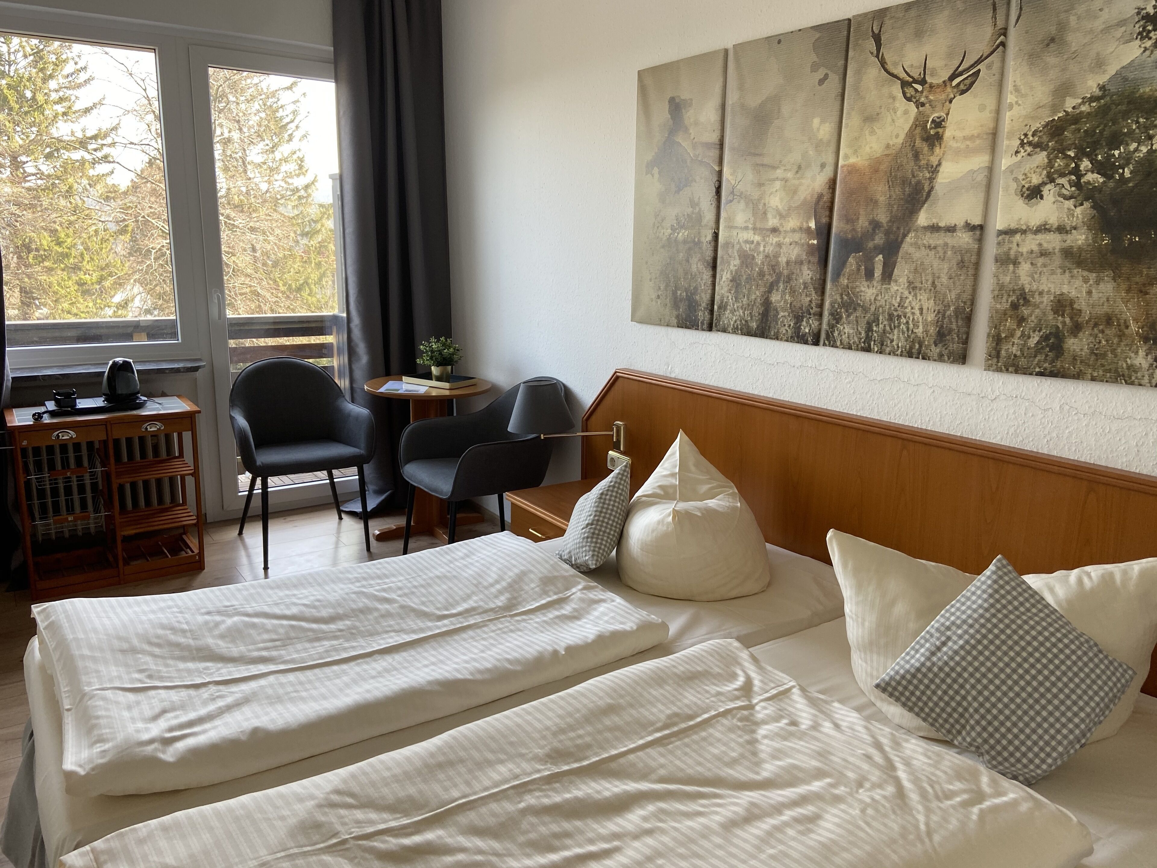 Una camera d'albergo con due letti, una scrivania, sedie e un'ampia finestra vista alberi.
