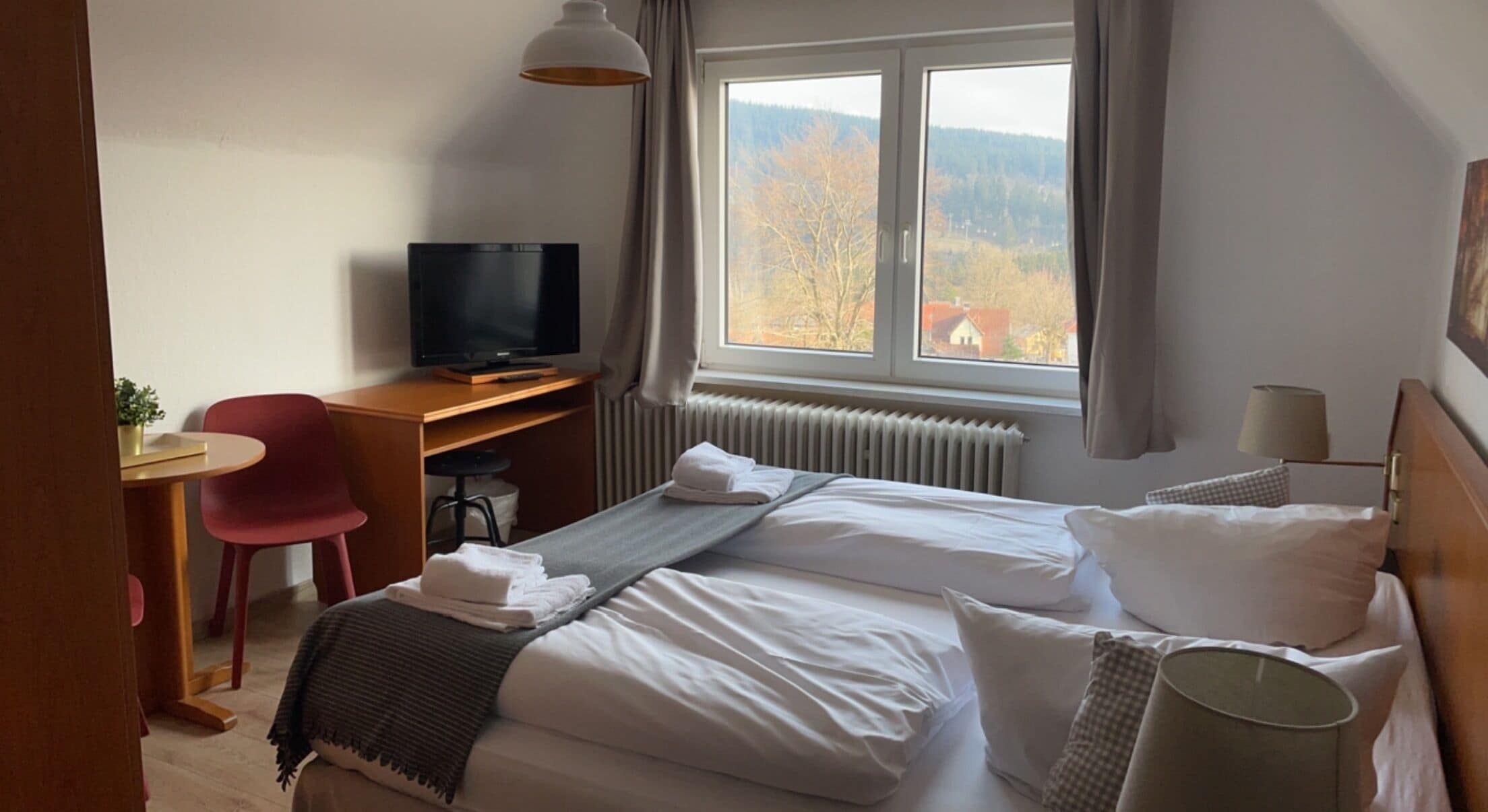 Et hotelværelse med to senge, et skrivebord med tv, en stol, et vindue med udsigt til træer og bygninger, samt en radiator.