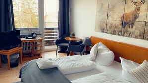 Quarto de hotel com uma cama, televisão, mesinha com abajur, cadeira e varanda com vista para árvores.
