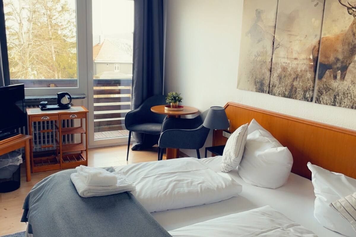 A hotel room with a bed, a TV, a small table with a lamp, a chair, and a balcony with a view of trees.
