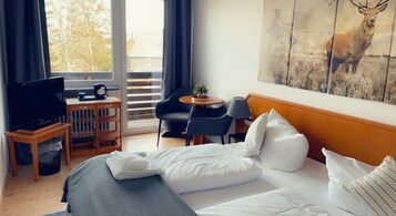 Quarto de hotel com uma cama, televisão, mesinha com abajur, cadeira e varanda com vista para árvores.