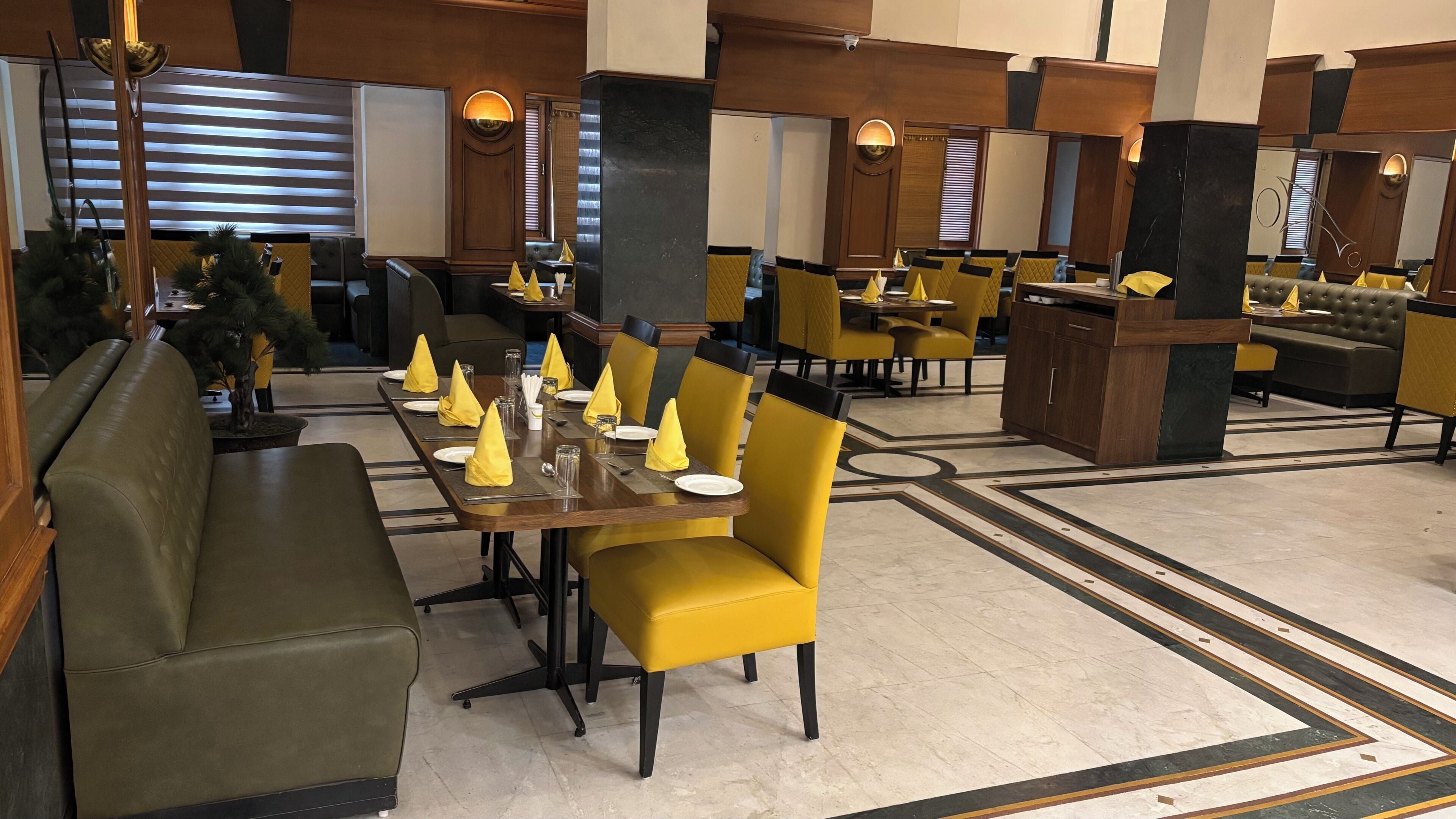 Um restaurante com várias mesas e cadeiras, algumas com guardanapos amarelos. Possui áreas de estar com almofadas e piso estampado.