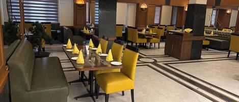 Um restaurante com várias mesas e cadeiras, algumas com guardanapos amarelos. Possui áreas de estar com almofadas e piso estampado.