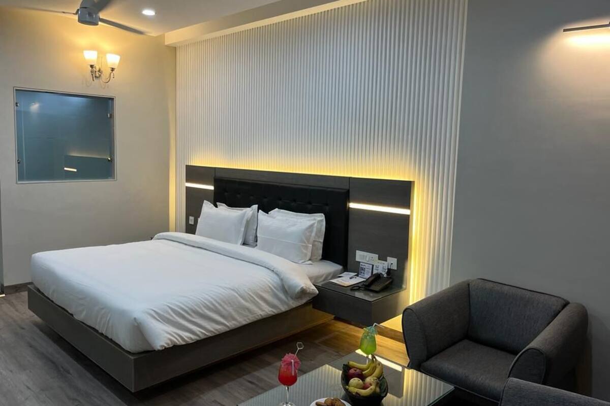 A hotel room with a bed, bedside table, chair, and a small table with a drink and fruit.