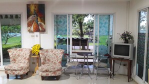 A living room with two floral armchairs, a small round table, a television on a stand, and a large window with a view of a garden. - RoomQuest Chonburi Phanat Nikhom (Phanat Nikhom)