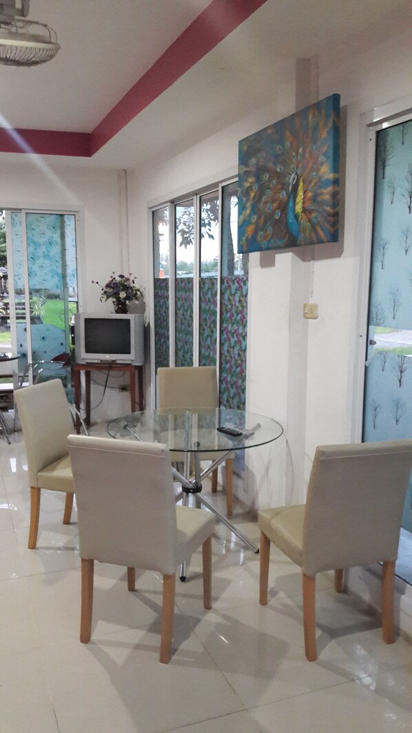 A dining area with a glass table, four chairs, a television on a stand, and a colorful painting on the wall. - RoomQuest Chonburi Phanat Nikhom (Phanat Nikhom)