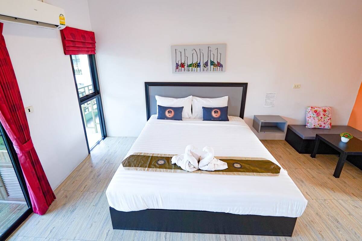 A hotel room with a large bed, a headboard, a window with red curtains, a wall-mounted air conditioner, a small table with a plant, and a wall art piece above the bed.