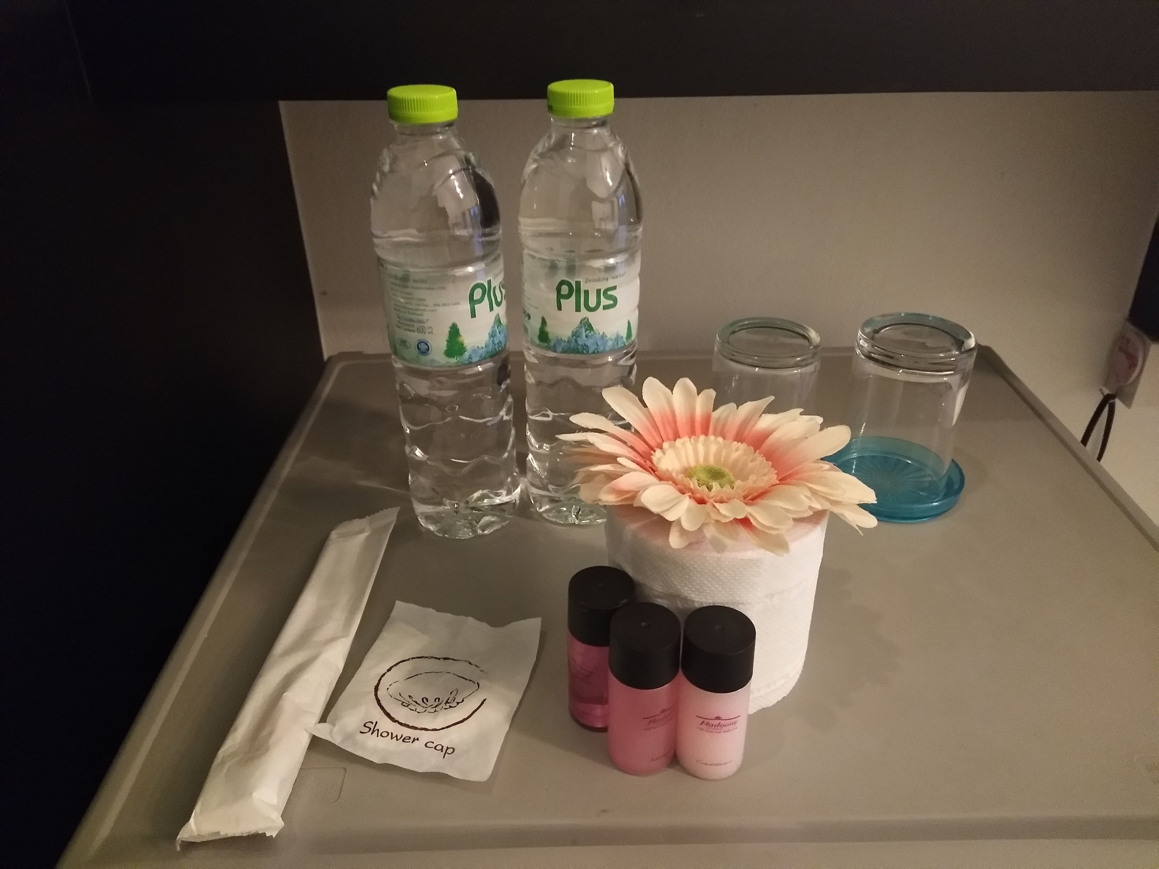 A tray with two bottles of Plus water, a tissue roll, a flower, and four bottles of shower cap.