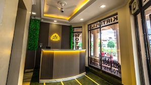 Zona de recepción de un hotel con diseño moderno, que incluye un mostrador de recepción, una pared verde y vistas a la calle a través de grandes ventanales.