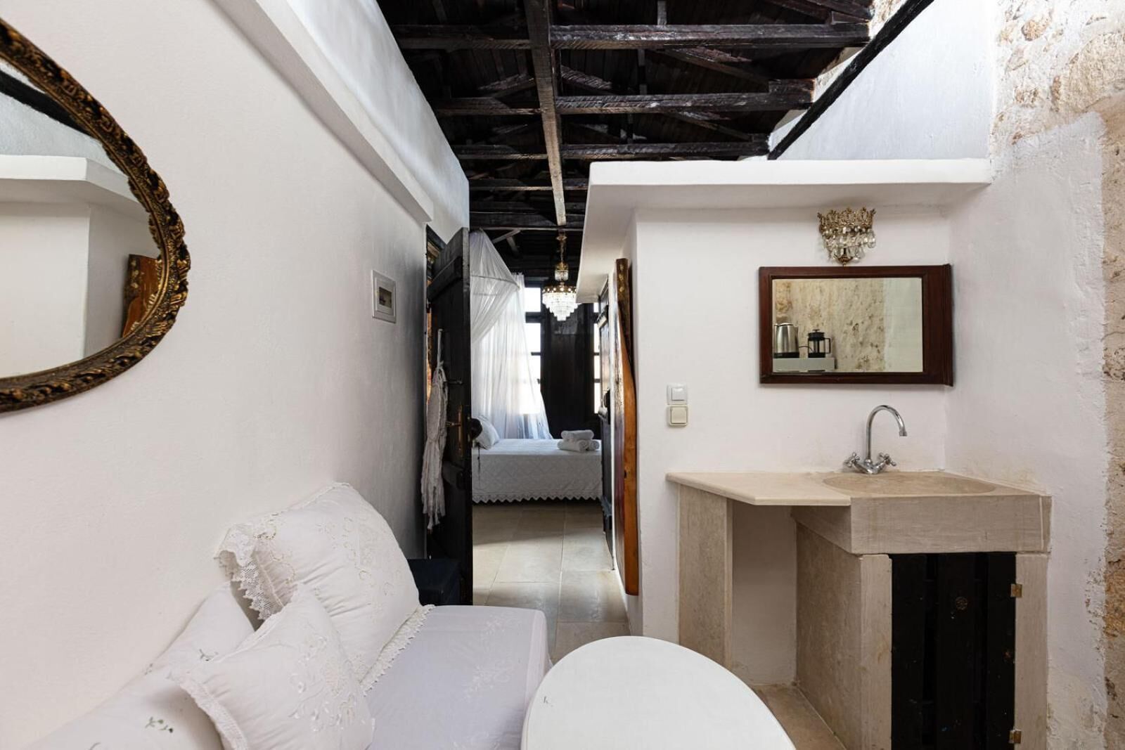 A narrow hallway leading to a bedroom with a bed, a white sofa, a bathroom with a sink and mirror, and a chandelier.