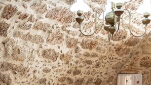 Ein Zimmer mit Steinwand, einem Bett mit weißer Bettwäsche, einem Tisch mit Stühlen, die mit leichtem Stoff bezogen sind, und einer Hängematte.