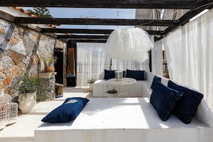 Un espacio al aire libre con luz solar, una pared de piedra, cojines blancos y un farolillo blanco.