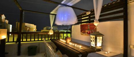 A covered outdoor seating area with a wooden bench, a table with candles, and a lantern.