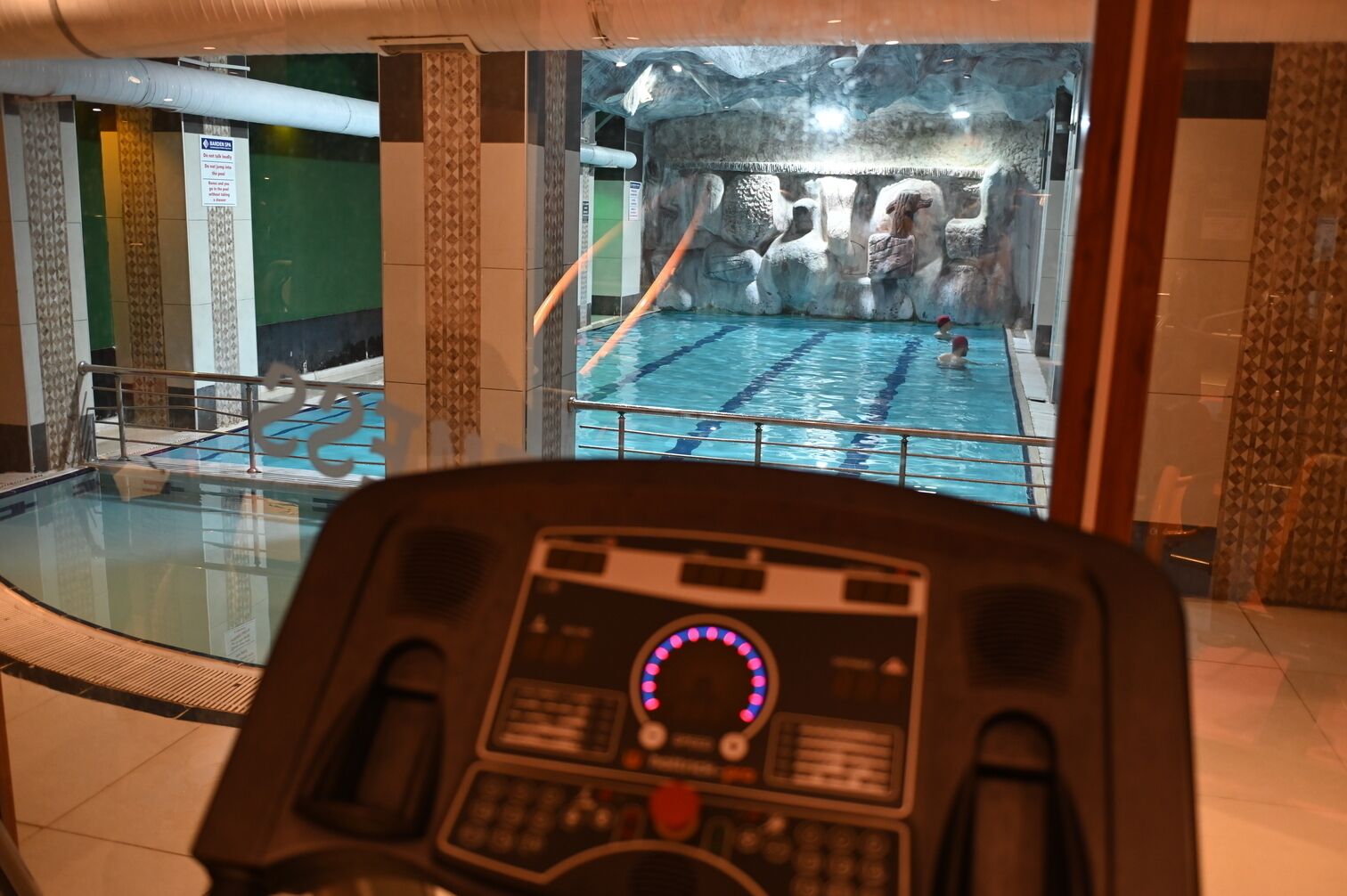 Una piscina interior con un fondo de pared rocosa, un panel de control en primer plano y un suelo de baldosas.
