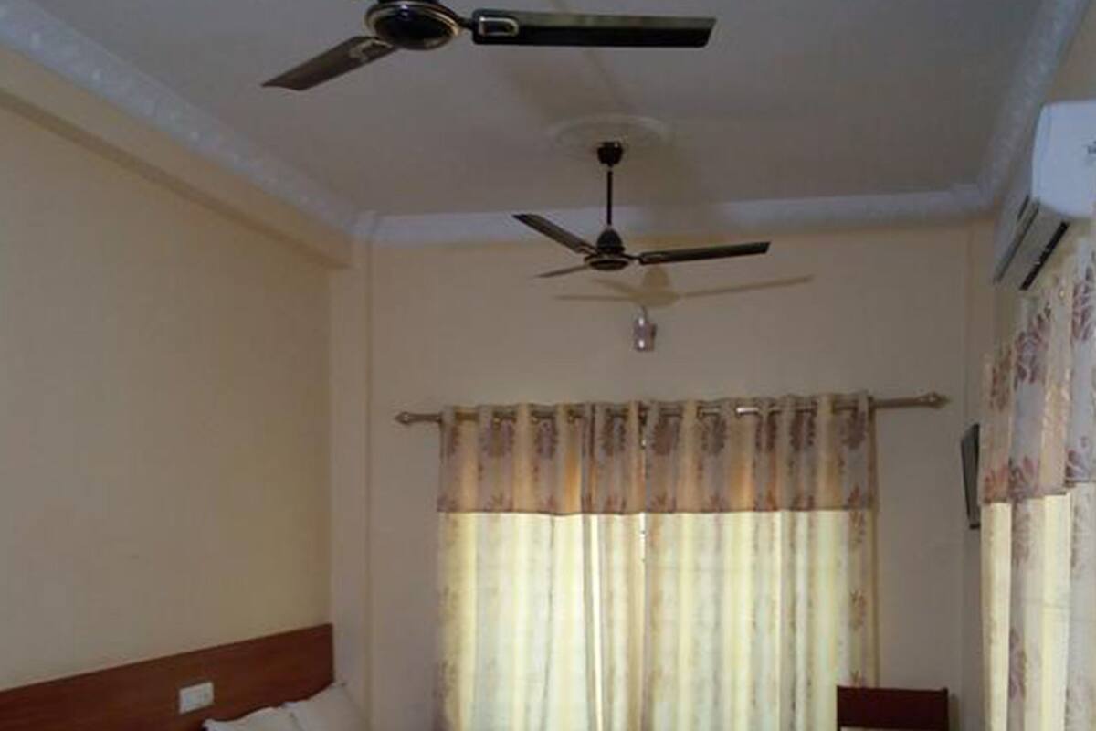 Quarto de hotel com duas camas, um ventilador de teto, uma janela com cortinas, uma mesa de cabeceira de madeira e uma cadeira.