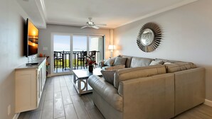 A modern living room with a sofa, a coffee table, and a TV. There is a balcony with a view and a ceiling fan.