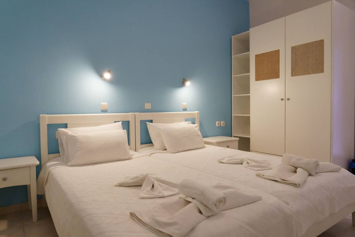 Ein ordentlich bezogenes Bett mit weißen Leinen und Kissen, ein weißer Nachttisch, ein Schrank mit durchbrochenen Lamellen und zwei Wandleuchten.