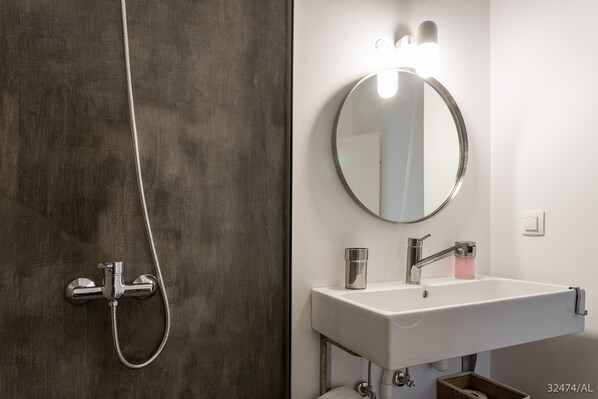 A bathroom with a modern shower, a white sink, and a round mirror with lights above.