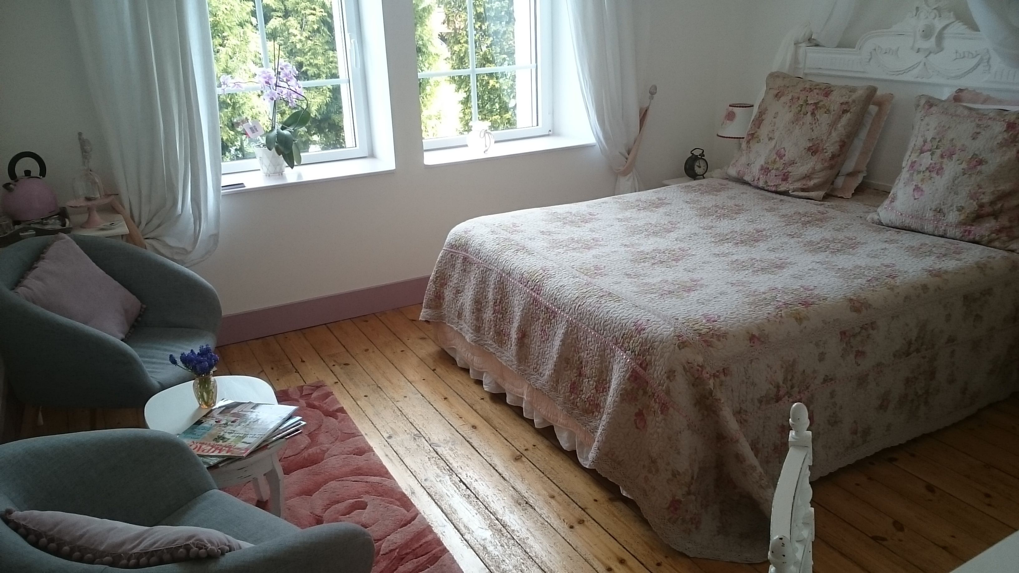 Un dormitorio con cama, una silla, una mesita con un jarrón y una ventana con cortinas.