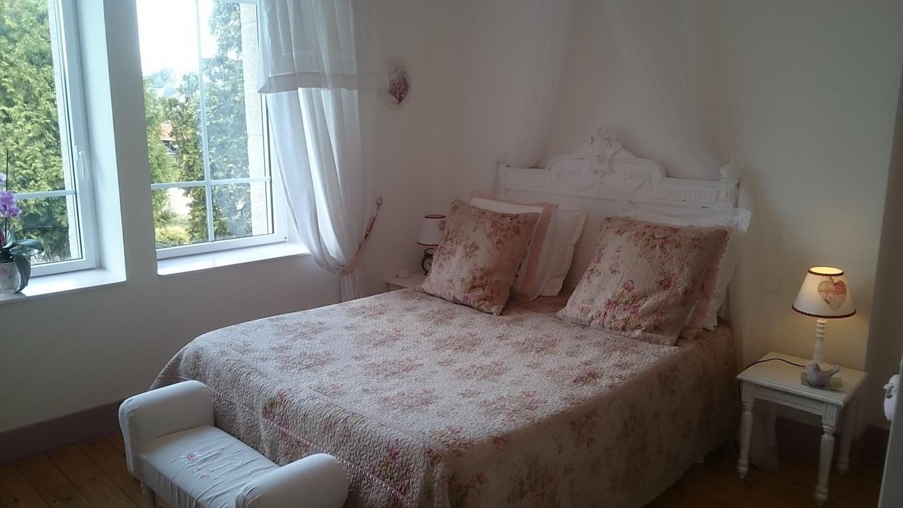Un dormitorio con cama, mesita de noche, lámpara, silla y ventana con cortinas.